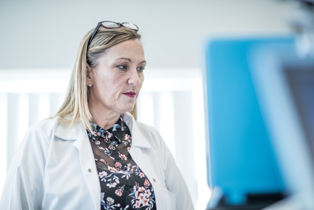 A photo of Nicola Warburton, wearing a white lab coat, looking towards a computer screen.